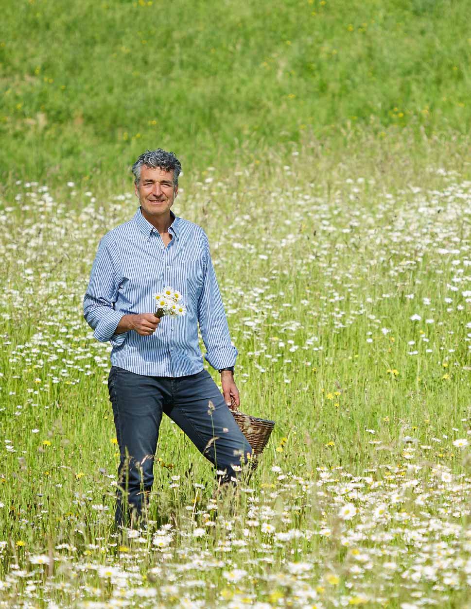 Mann im blau gestreiften Hemd steht mit Korb und weißen Blüten in der Hand in einer Blumenwiese