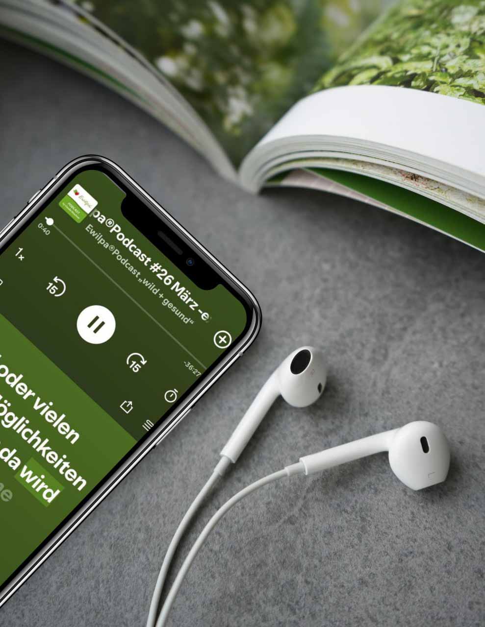 Smartphone spielt die Folge „Ewilpa®Podcast #26 März“, daneben Ohrhörer und ein aufgeschlagenes Magazin