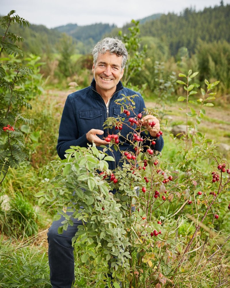 Dr. Markus Strauß steht lächelnd an einem Hagebuttenstrauch voller roter Früchte vor bewaldeten Hügeln