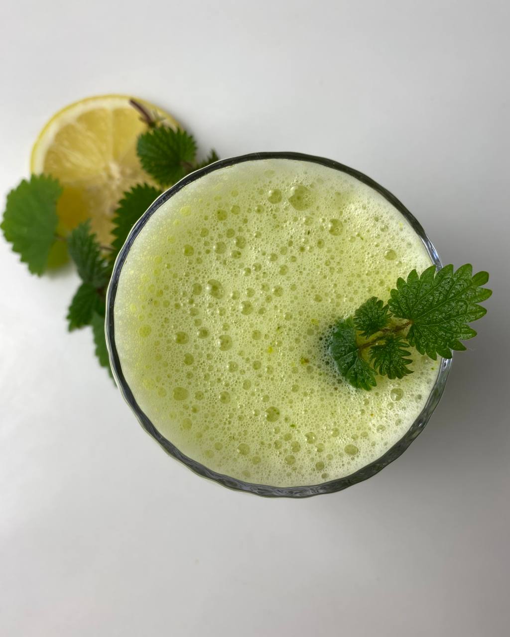 Schaumiger grüner Brennnessel-Smoothie im Glas, daneben Blätter und eine Zitronenscheibe