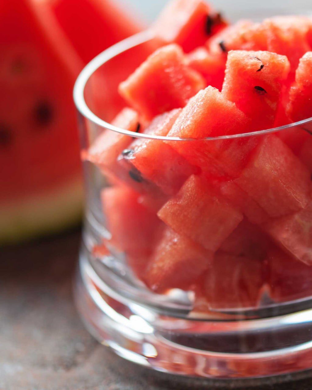 Glas mit gewürfeltem rotem Melonenfruchtfleisch, dahinter Melonenspalten