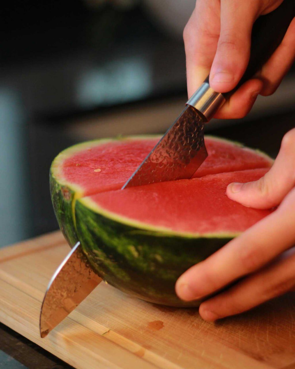 Hände schneiden mit einem Messer eine halbierte Melone mit rotem Fruchtfleisch auf einem Holzbrett