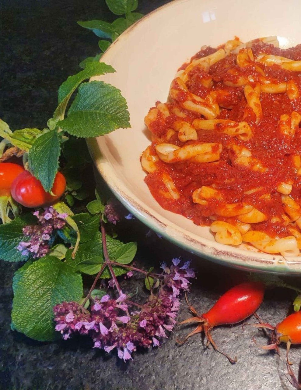 Pasta mit roter Hagebutten-Soße in einer Schale, daneben Hagebutten und Oreganoblüten