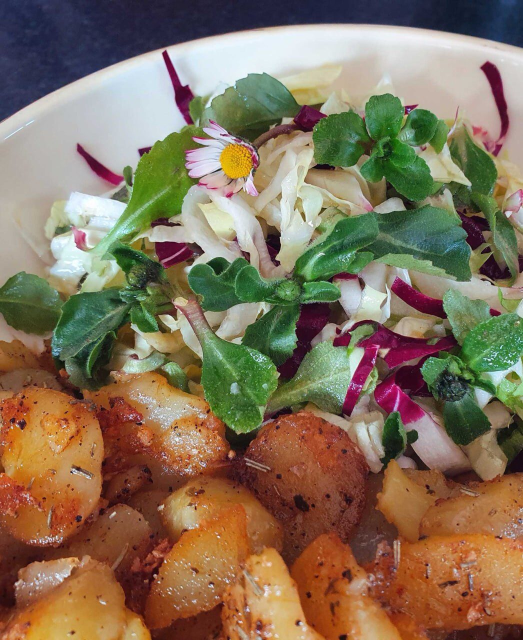 Schüssel mit Bratkartoffeln und wildem Blattsalat, obenauf eine Gänseblümchen-Blüte
