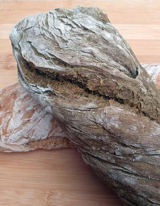 Zwei dunkle, aufgerissene Brotlaibe aus Laubmehl und Wurzeln auf einem Holzbrett