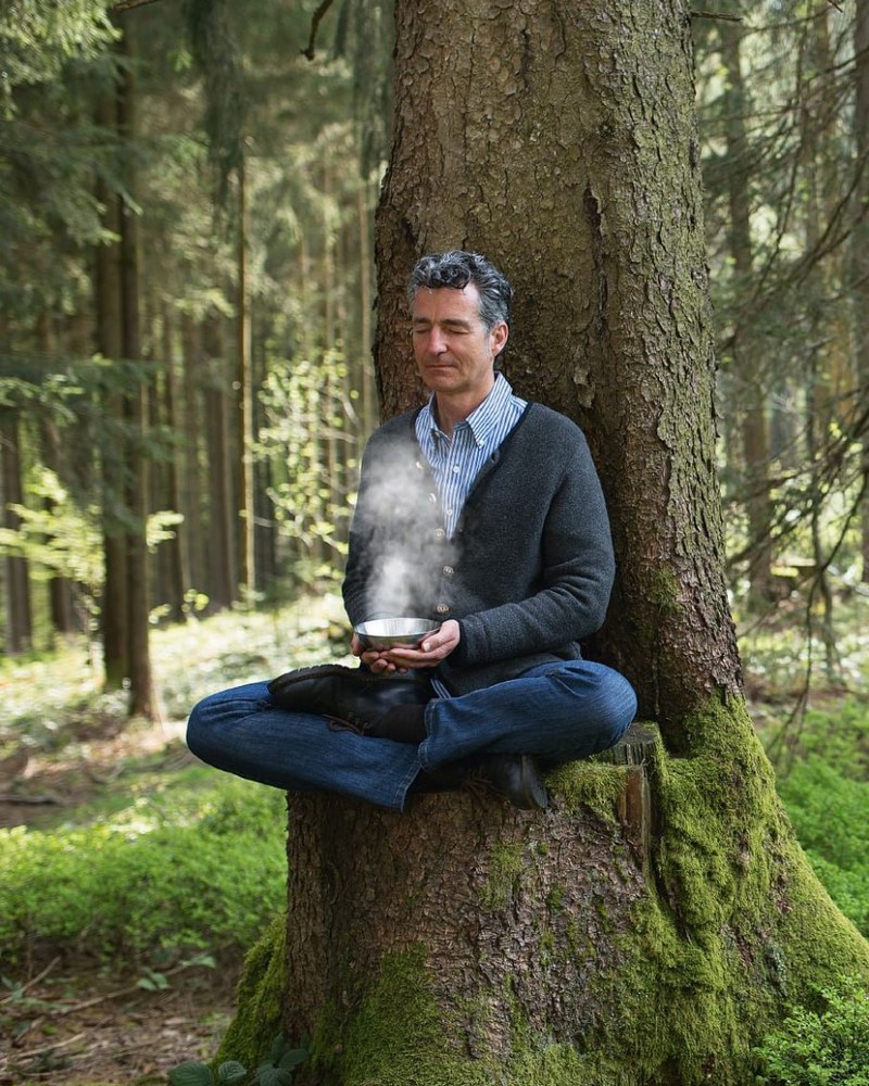Dr. Markus Strauß sitzt mit einer dampfenden Schale in den Händen auf einem bemoosten Baumstumpf im Nadelwald